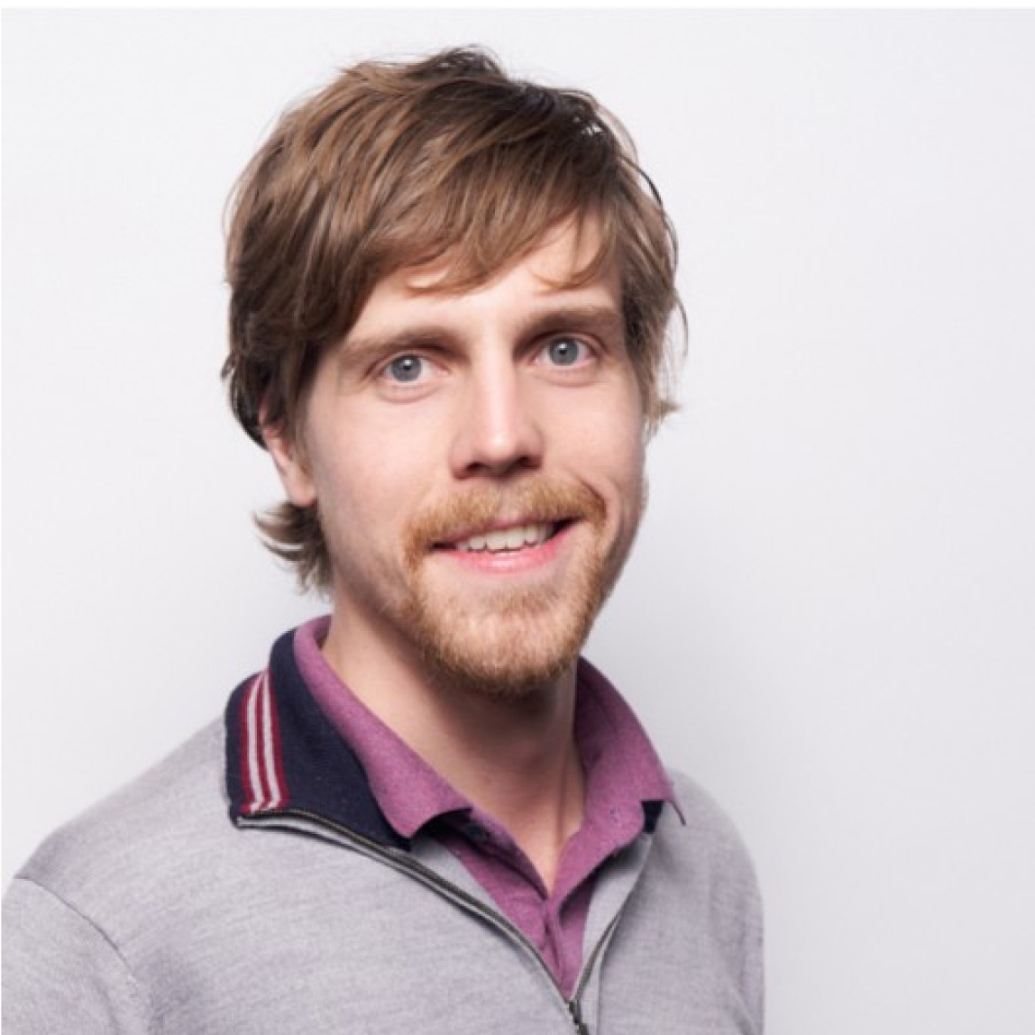 Man with sweater and purple collared shirt in front of a white background.